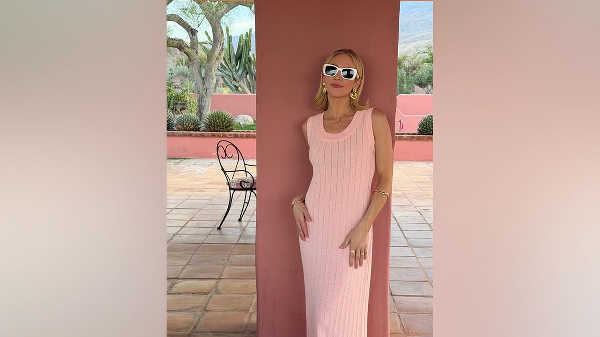 Brittany Snow leaning against a pink wall wearing a sleeveless pink knit dress and large gold earrings.