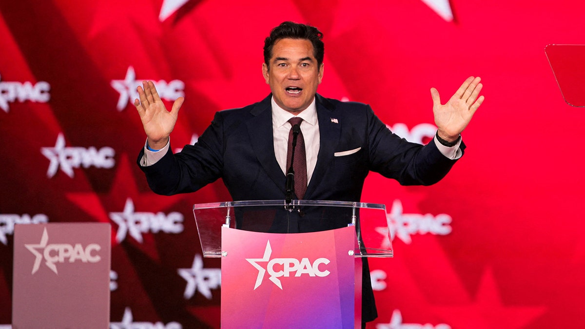 Wide shot of Dean Cain at a podium on a CPAC stage, gesturing with arms outstretched beneath a large CPAC logo on a red backdrop.