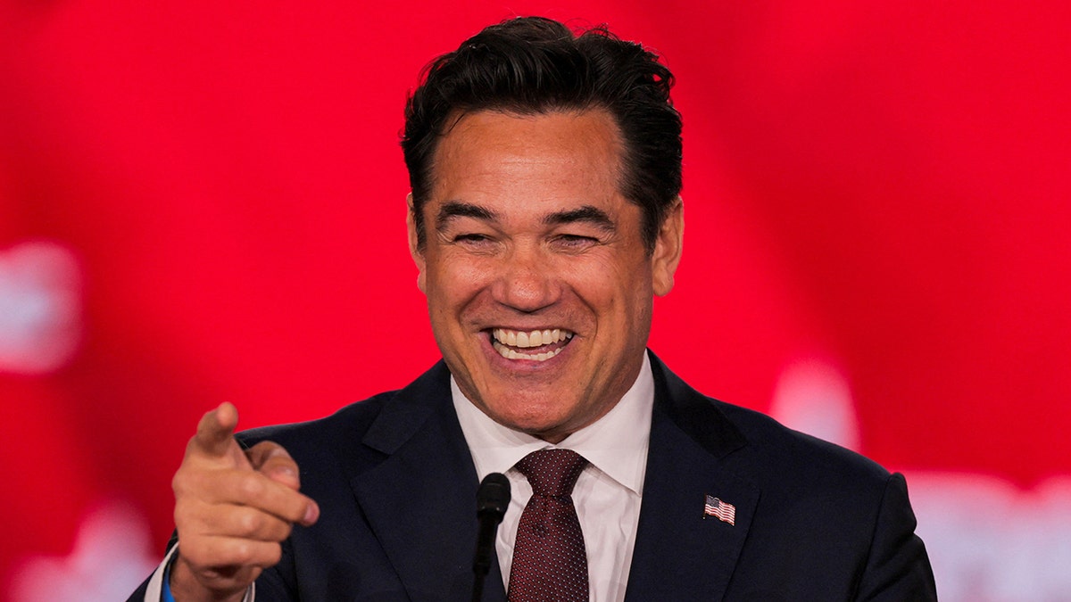 Close-up of Dean Cain smiling at a podium and pointing toward the audience, wearing a navy suit and burgundy tie with a CPAC backdrop behind him.
