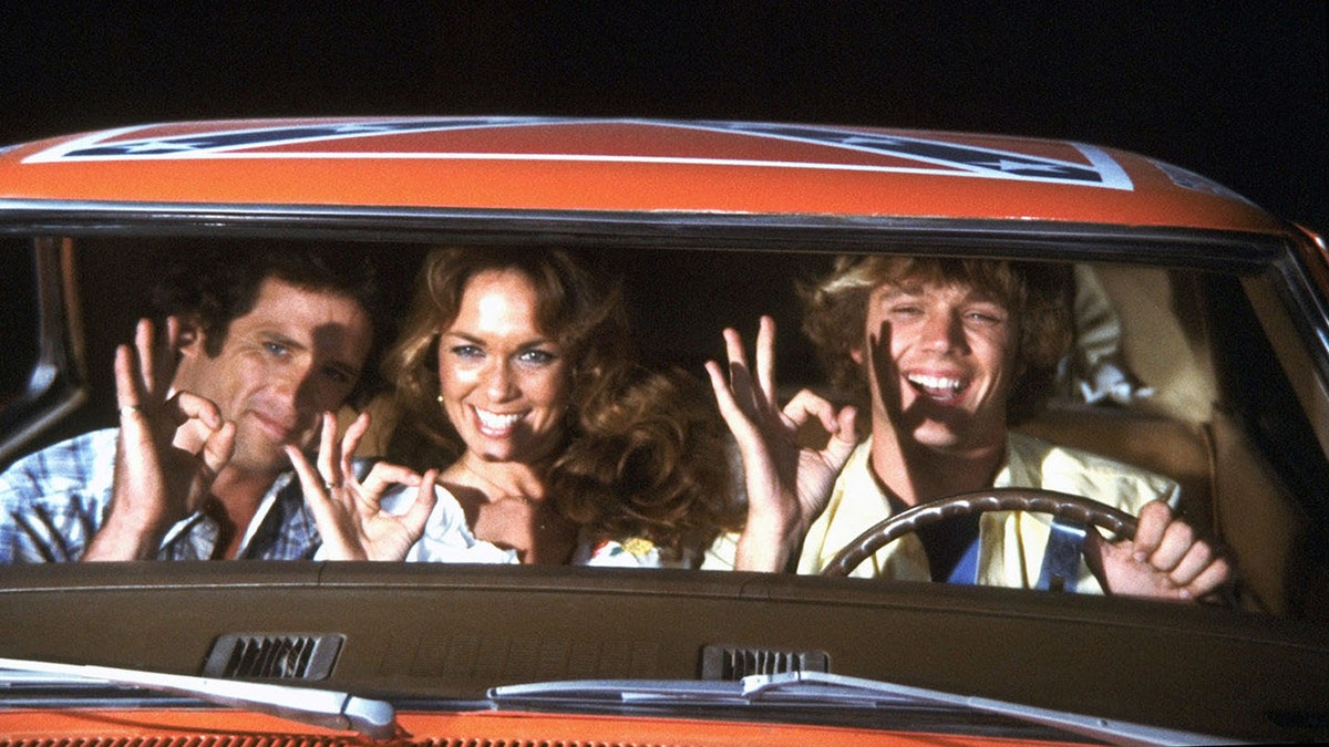 The cast of The Dukes of Hazzard inside the General Lee giving an OK sign.
