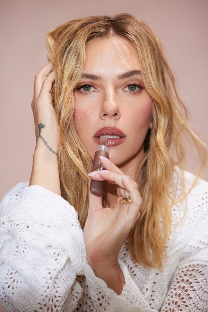 A woman with blonde hair and green eyes applies a clear gloss to her lips.