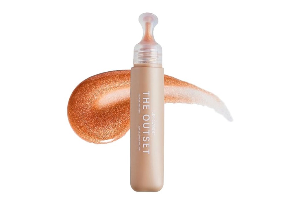 The Outset Lip Oasis Glossy Treatment in a beige tube with a clear applicator, next to a swatch of shimmering, peachy-gold lip gloss.