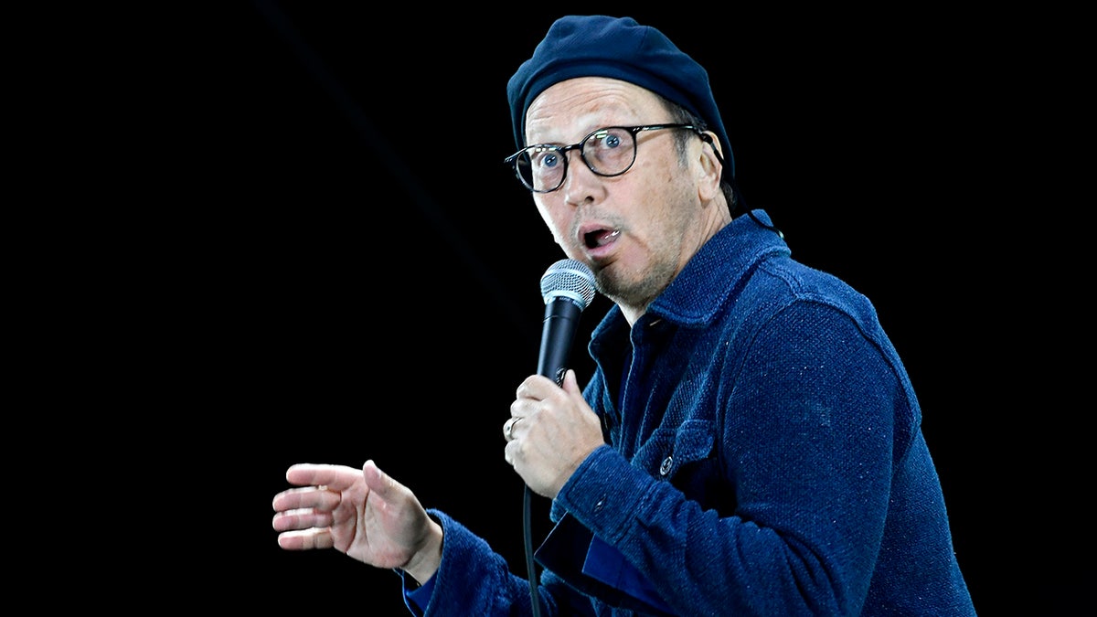 Rob Schneider in a blue jacket and backwards paperboy hat has his mouth ajar while on stage