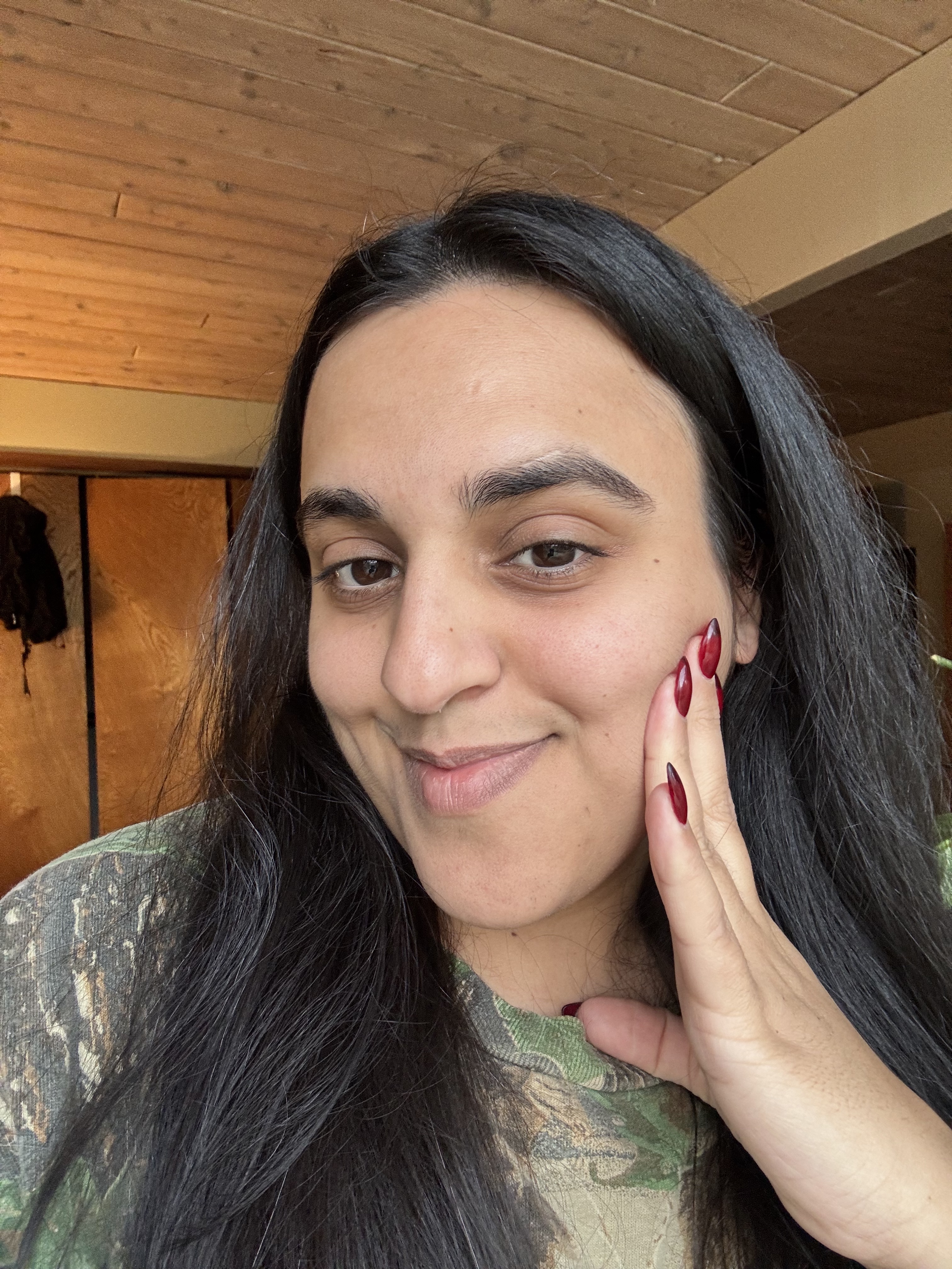 A woman with long dark hair, wearing a camouflage shirt, smiles while touching her cheek.