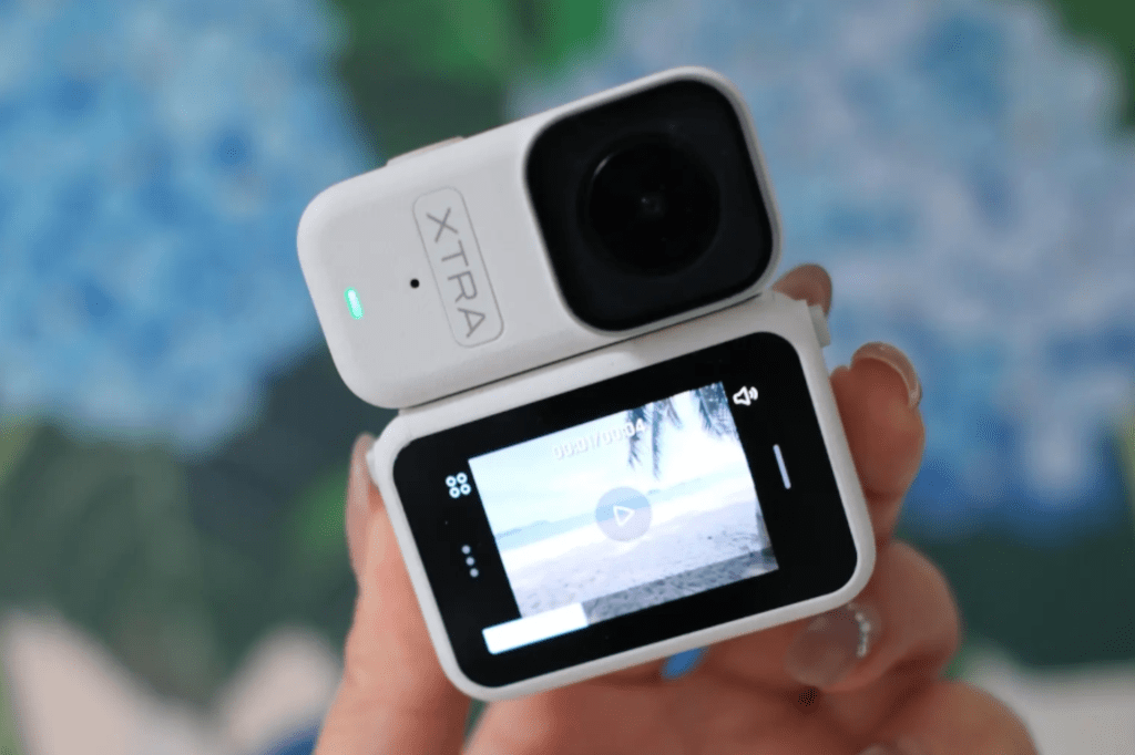 A person holding a white Xtra-Atto action camera, displaying a beach scene on its screen.