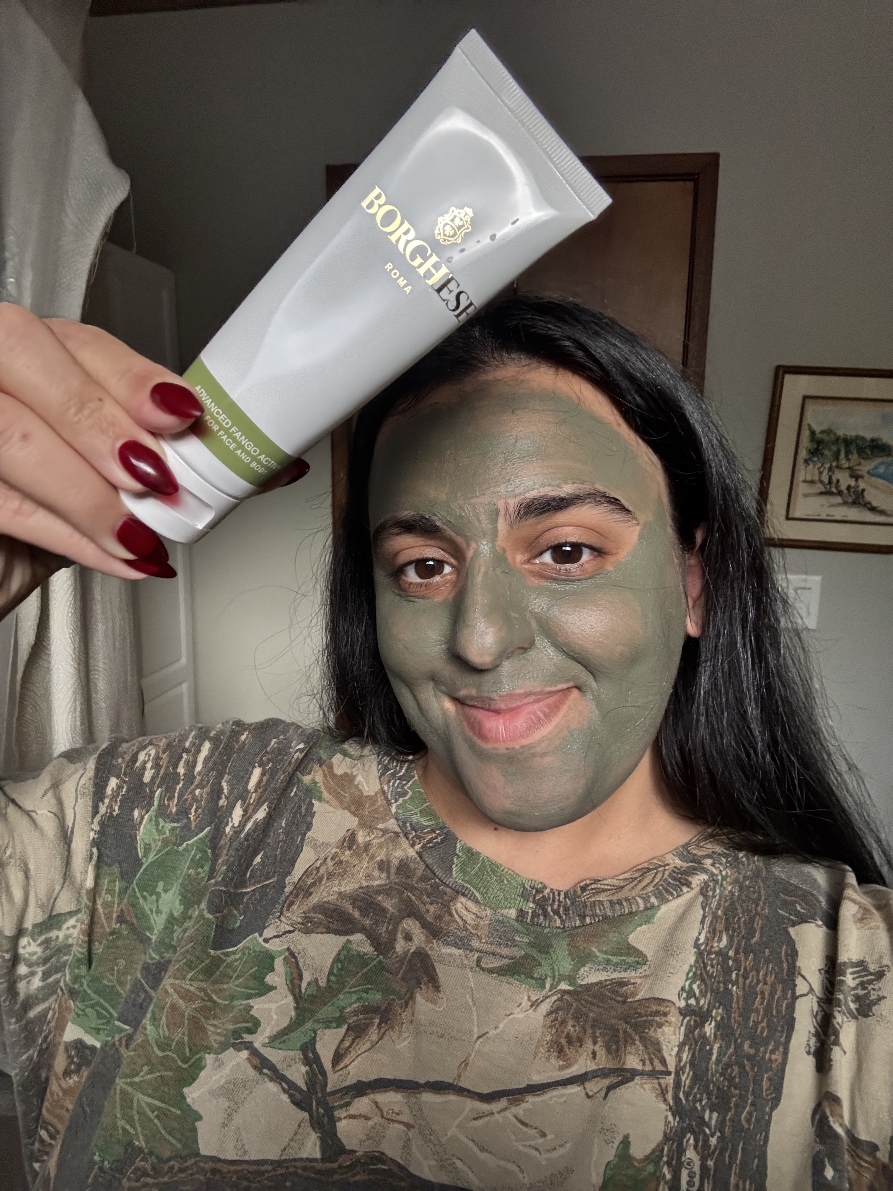 A woman with a green clay mask on her face holds up a tube of Borghese skincare.