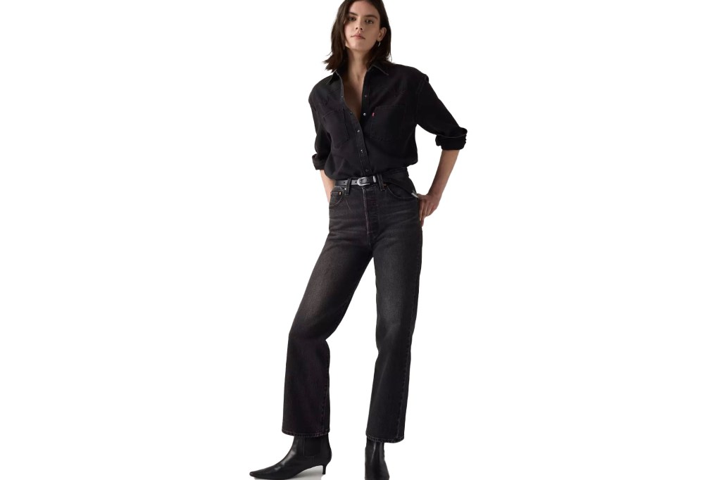 A woman posing in a black button-up shirt, black belt, and faded black jeans.