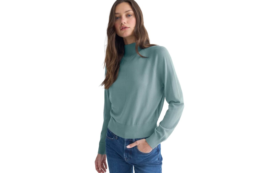 A woman with long brown hair wearing a long-sleeved teal sweater and blue jeans.