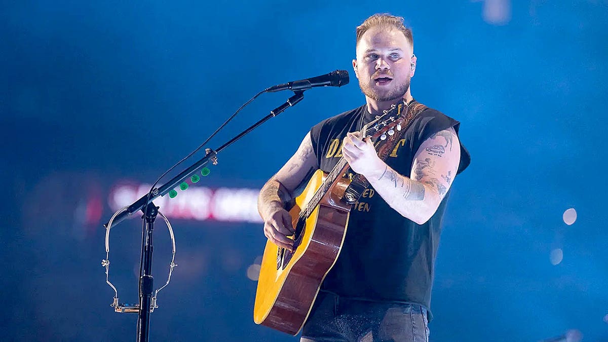 Zach Bryan playing guitar while standing in front of a microphone during a concert