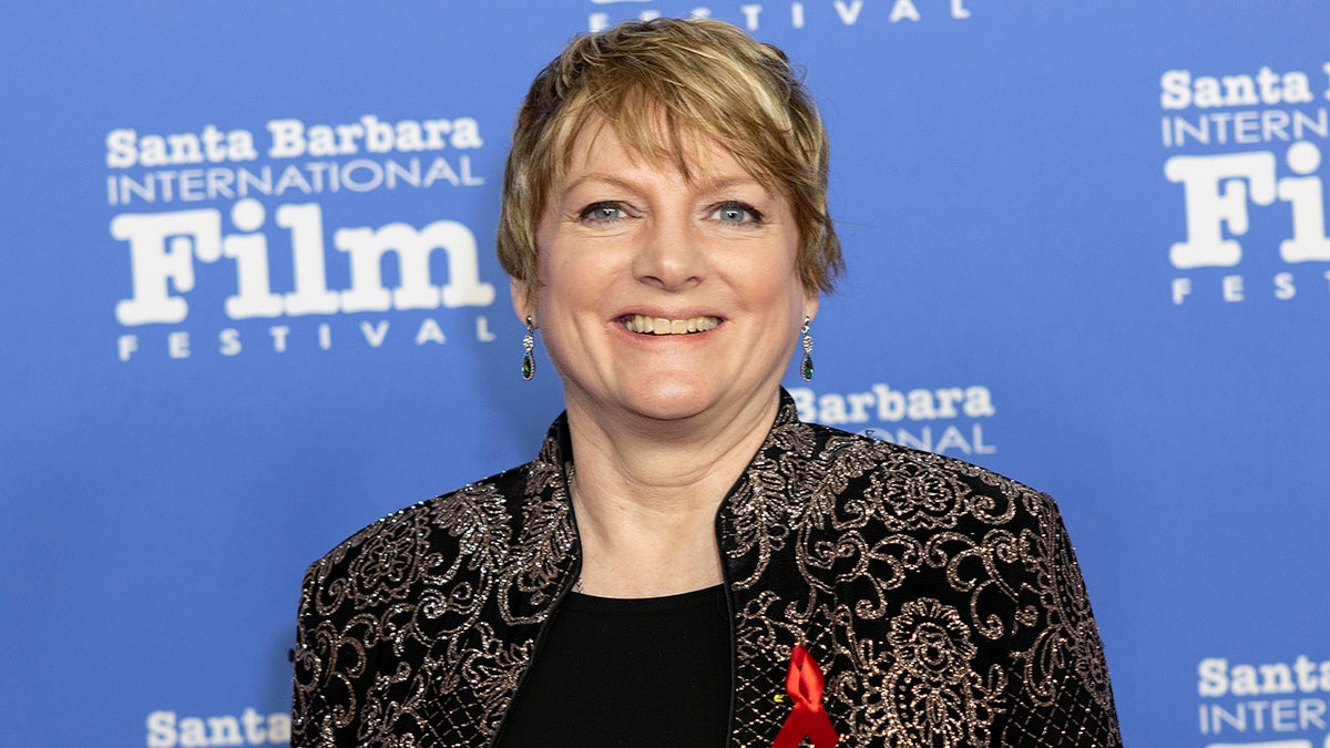 Nellie Olesone smiling wearing a black blouse and a patterned gold blazer
