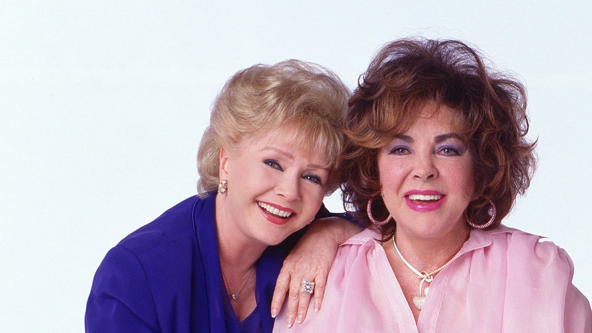Debbie Reynolds wearing blue blouse smiling leaning on Elizabeth Taylor wearing pink blouse smiling