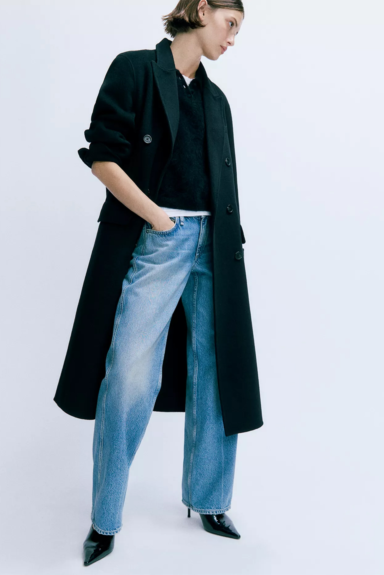 A model wearing a black coat, a black sweater, low-rise jeans, and black pointed-toe heels.
