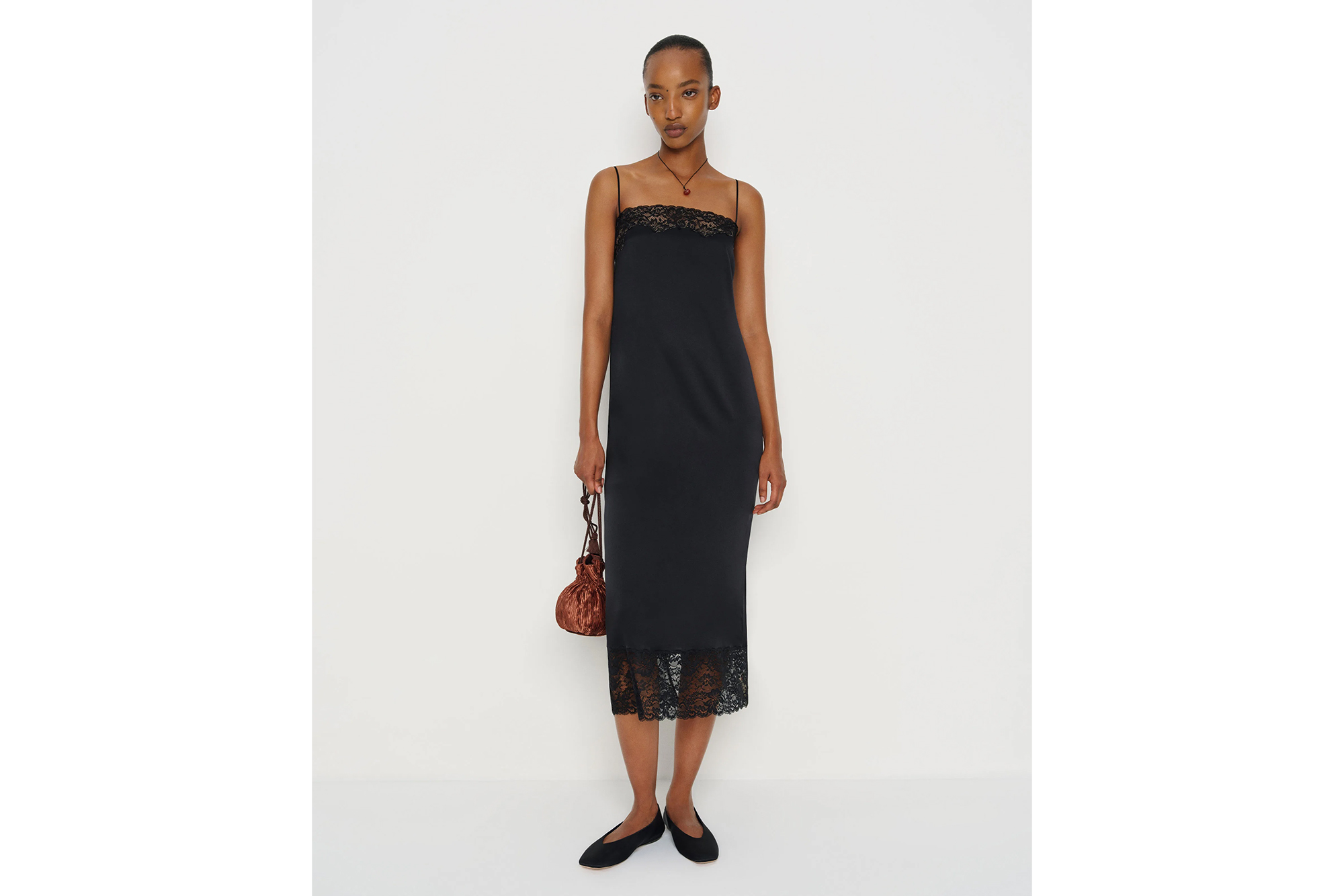 A model poses in a black midi-length slip dress with lace trim, holding a small brown velvet drawstring bag.