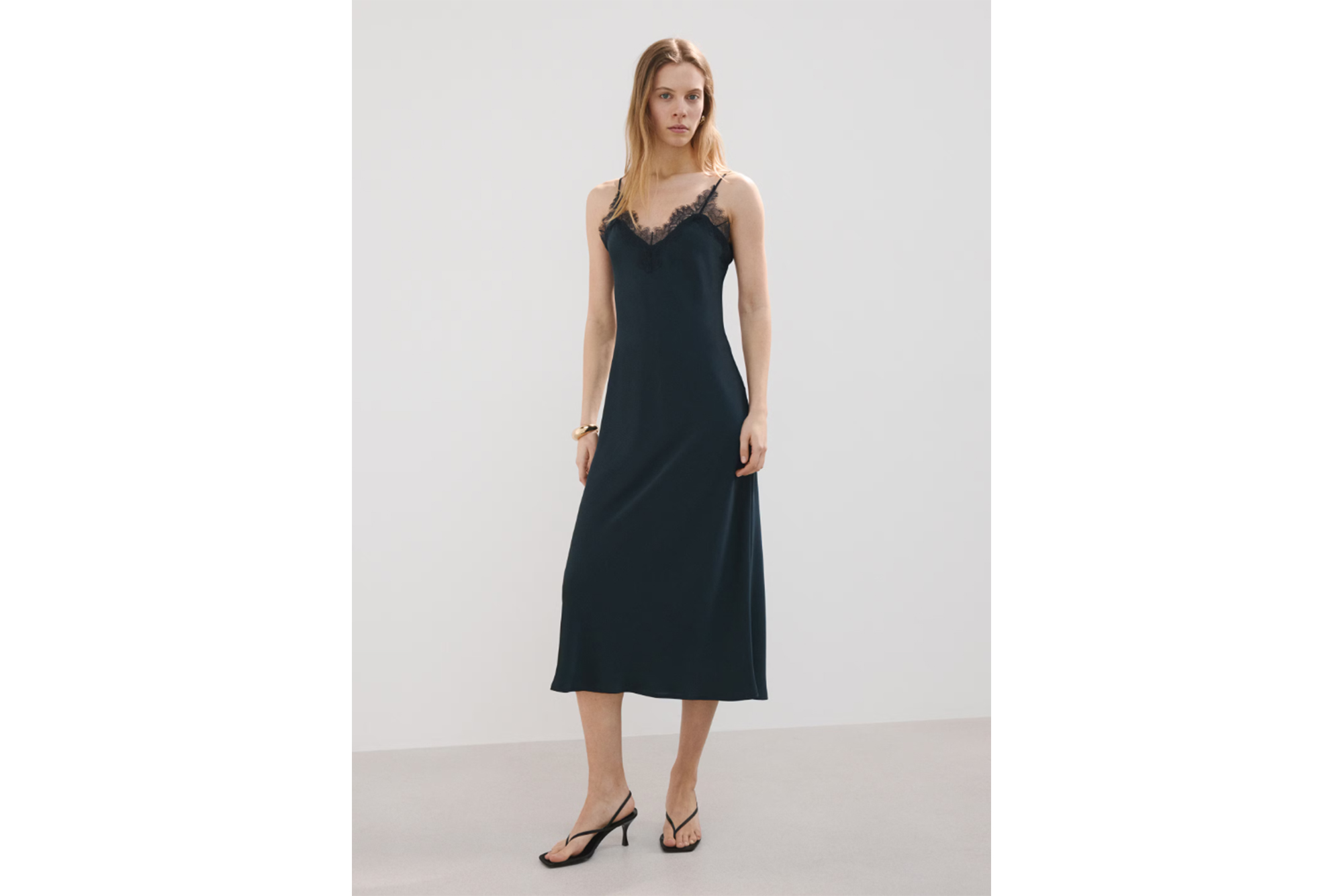 Model wearing a black slip dress with lace trim and black kitten-heel thong sandals.