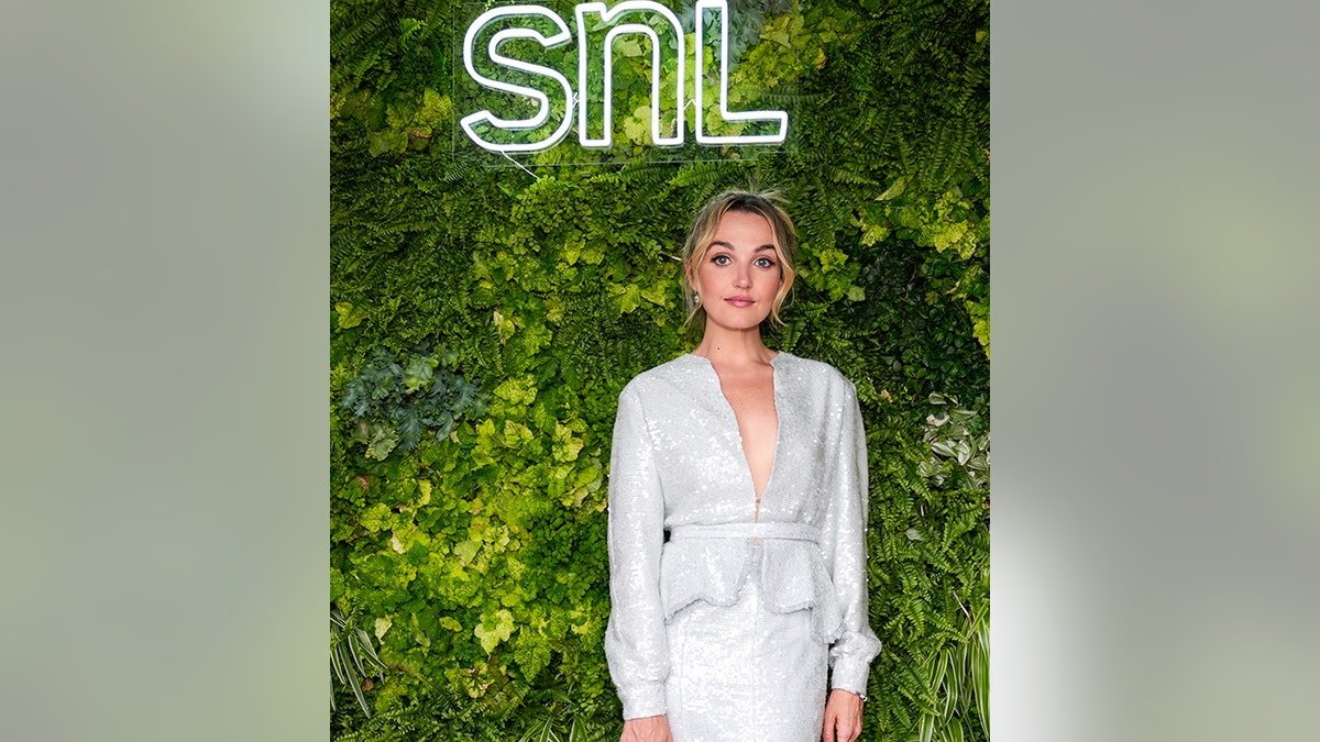 Chloe Fineman wearing sparkly blazer and skirt standing in front of foliage and SNL sign