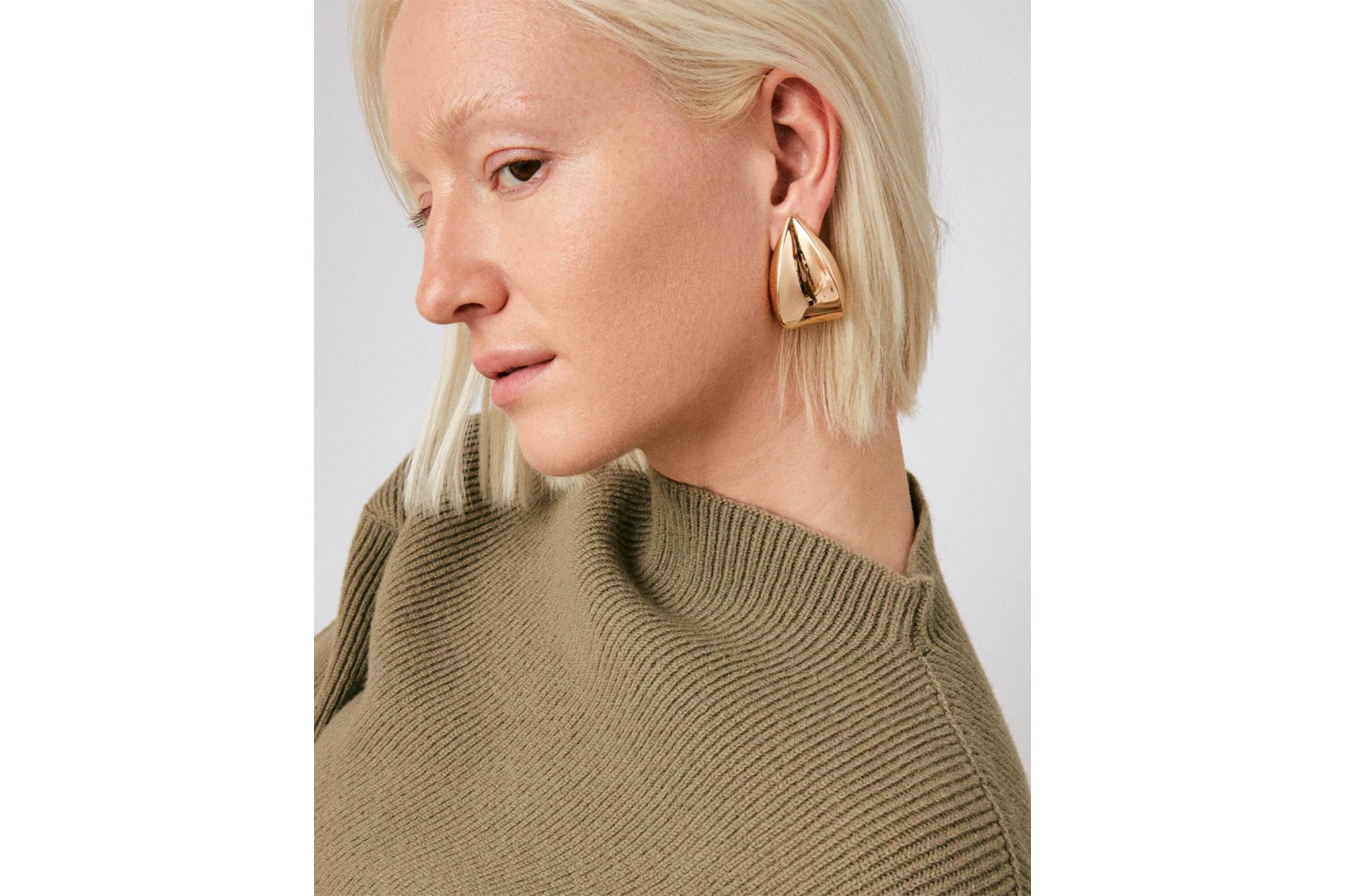 A woman with short blonde hair wearing a ribbed olive green sweater and a large triangular gold earring.