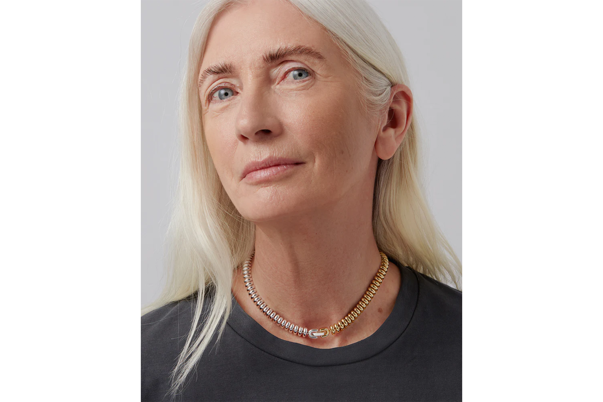 A woman with long white hair and a grey t-shirt modeling a gold-colored choker necklace with a rectangular clasp.