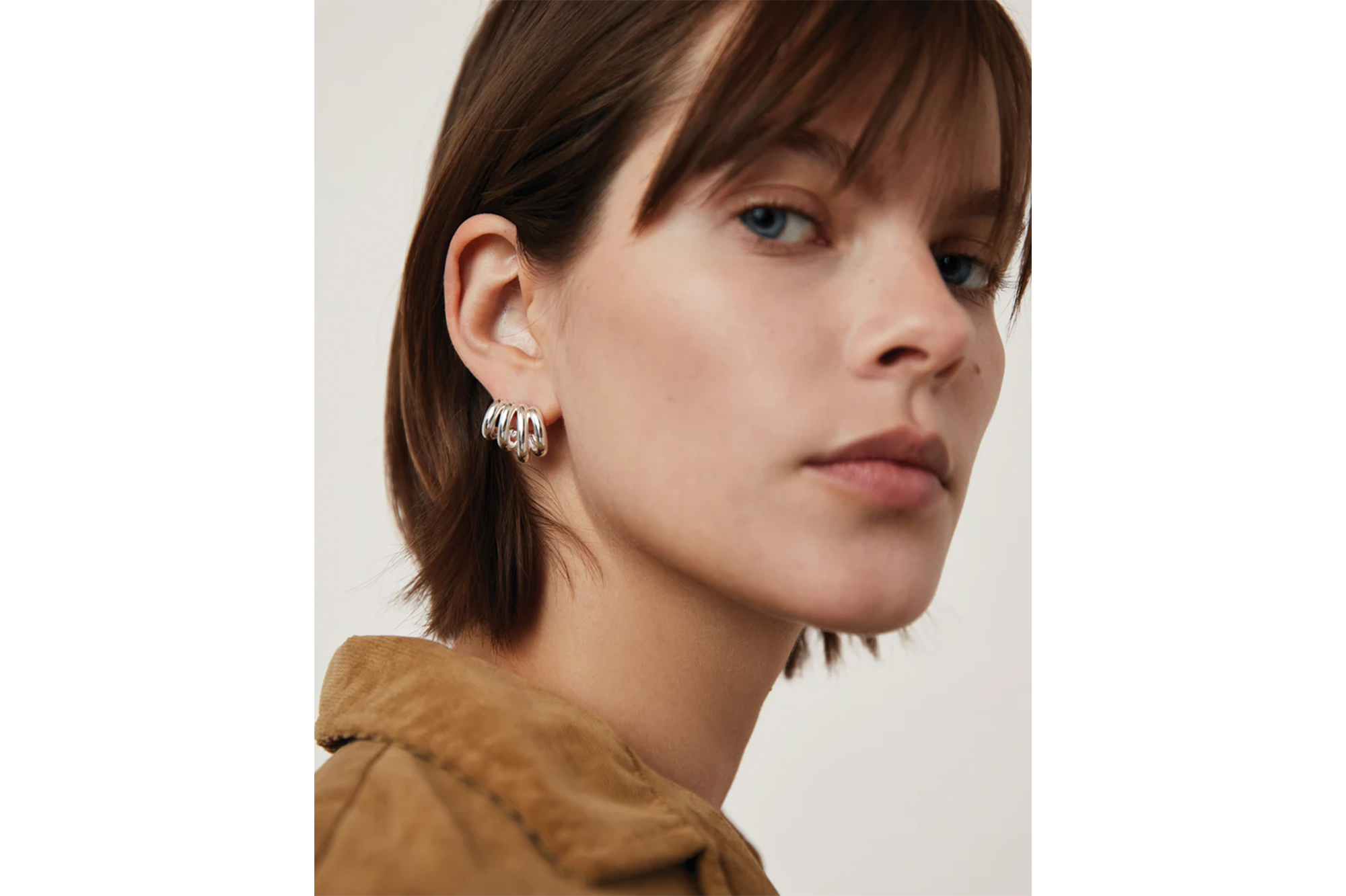 A model with short brown hair, blue eyes, and a brown corduroy jacket wears silver five-ring spiral stud earrings.