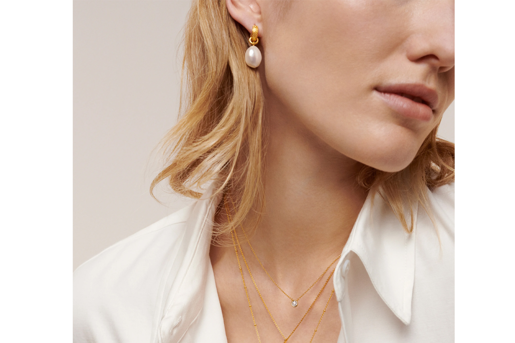 A person wearing a single baroque pearl drop earring and two layered gold chain necklaces with small pendants.