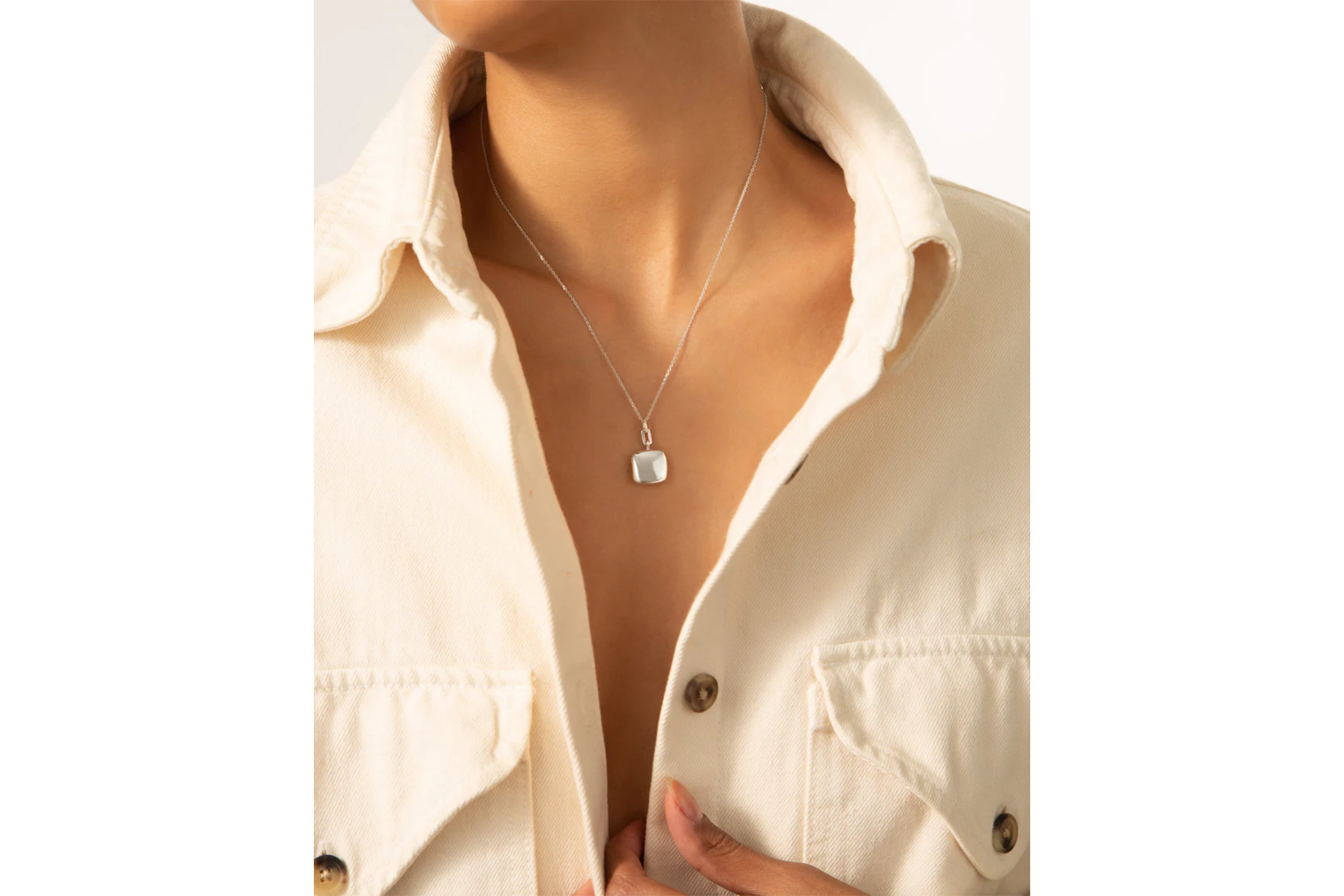 A person wearing a silver square pendant necklace over a cream collared shirt.