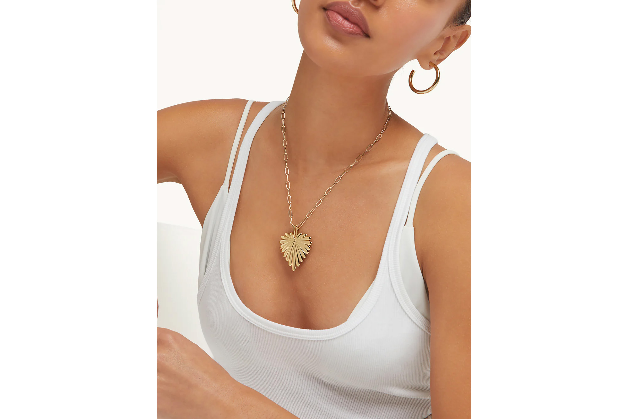 A model wearing a white tank top, gold hoop earrings, and a gold necklace with a large heart-shaped pendant.