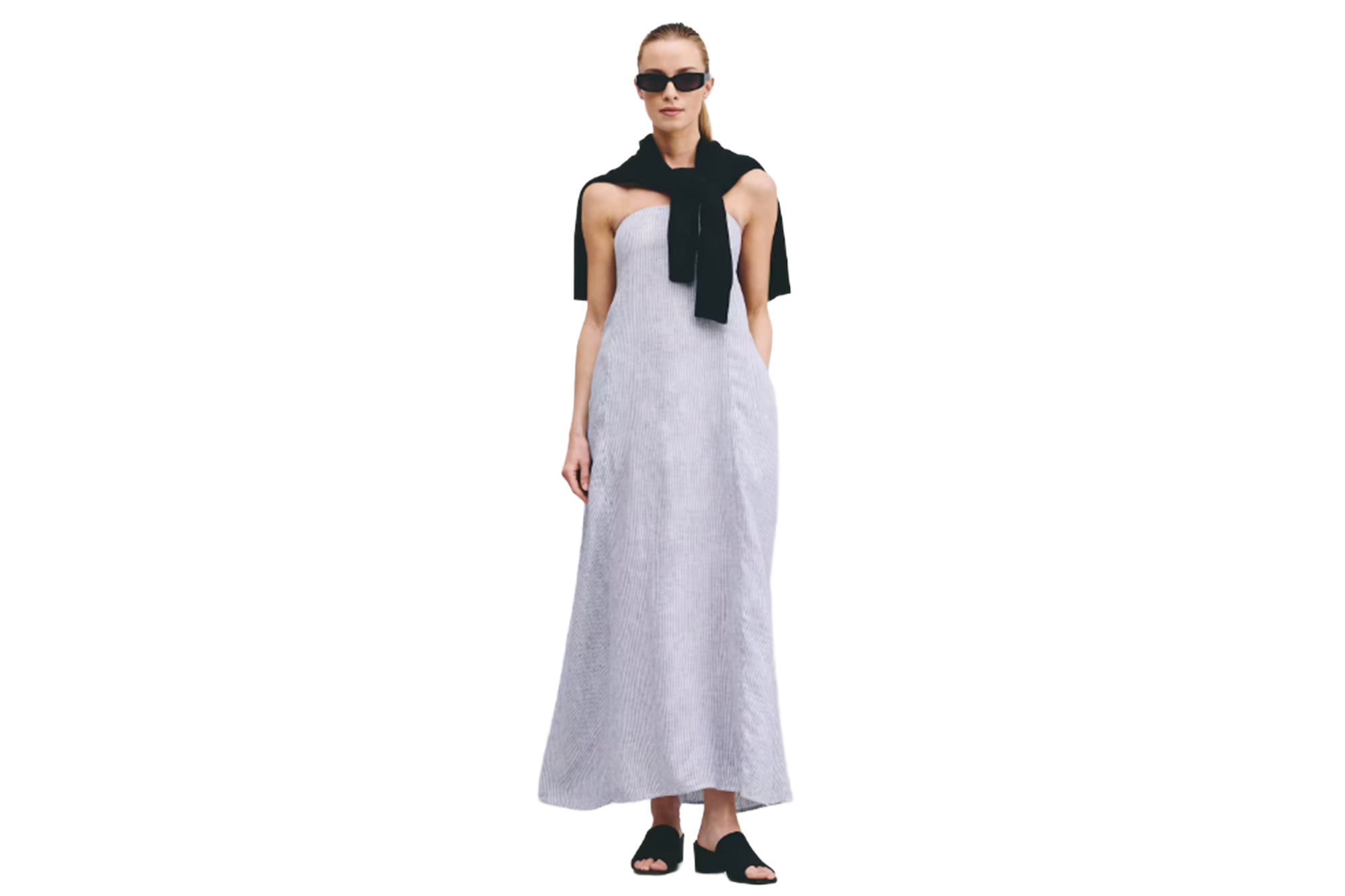 Woman in a light blue striped strapless maxi dress, black sweater draped over her shoulders, black sunglasses, and black open-toe flat shoes.