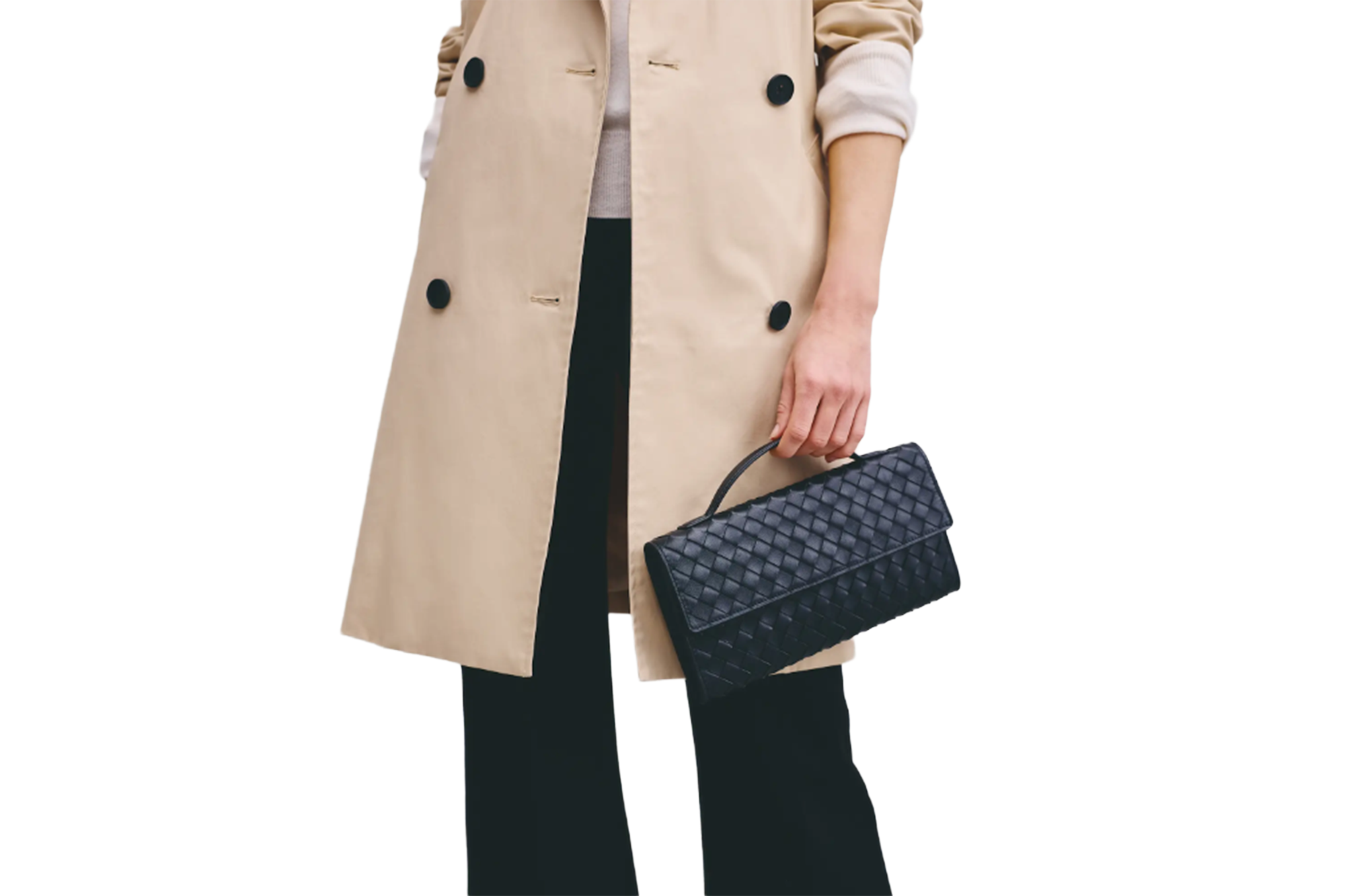 A person in a tan coat and black pants holding a black woven clutch bag.