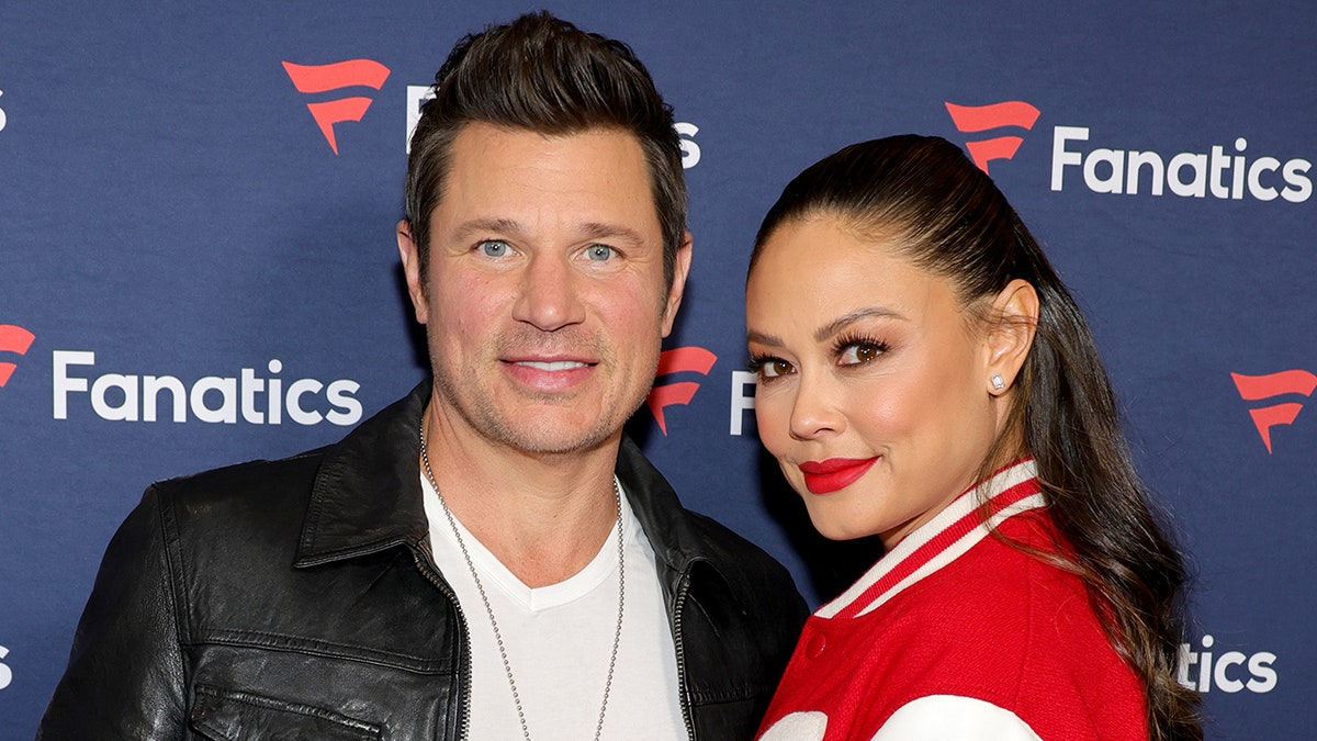Nick Lachey smiling in a leather jacket with wife Vanessa Lachey in a red letterman jacket at a party