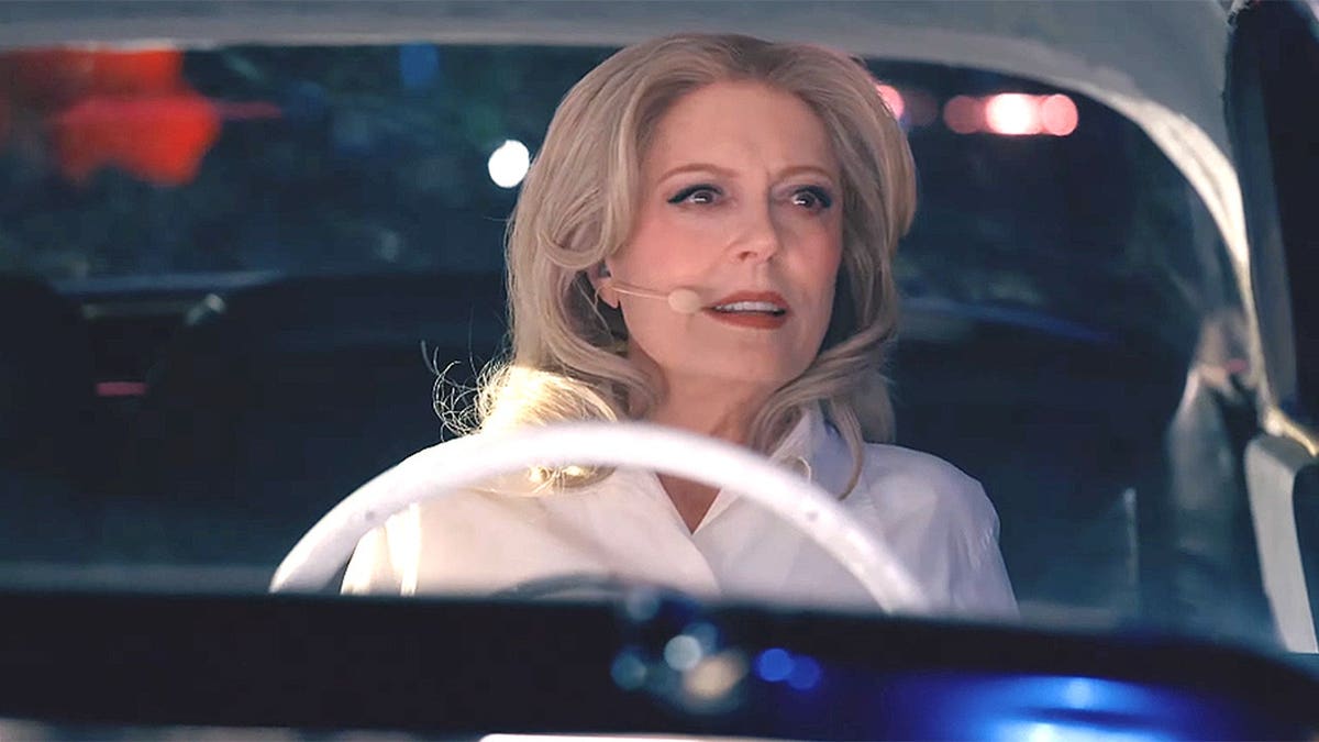 Susan Sarandon driving a vintage car, wearing a headset microphone and looking ahead with a focused expression.
