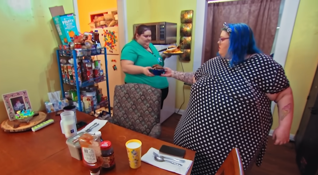 Two women in a messy kitchen. One, with blue hair and a polka dot dress, takes food from the other.