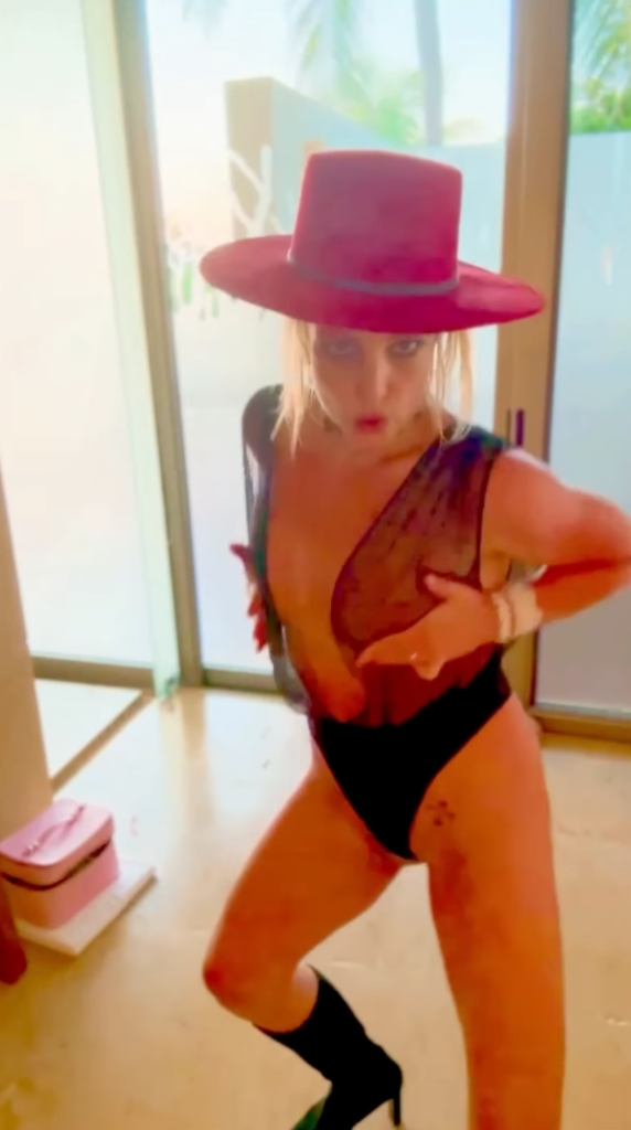 Britney Spears in a red hat, sheer black bodysuit, and black boots.