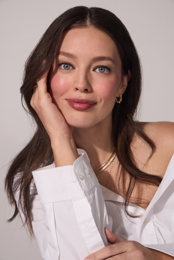 A woman with dark hair and blue eyes smiles while wearing a white shirt and gold jewelry.