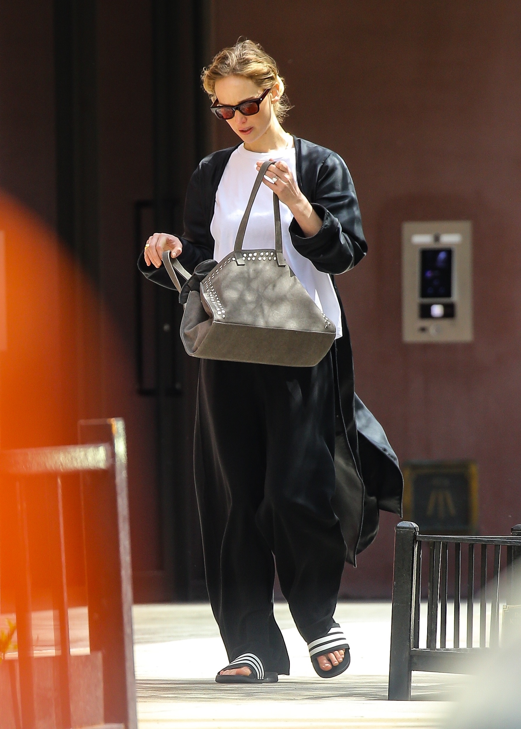 Jennifer Lawrence walking in a black cardigan, white tee, black pants, Adidas sandals, and a gray bag.