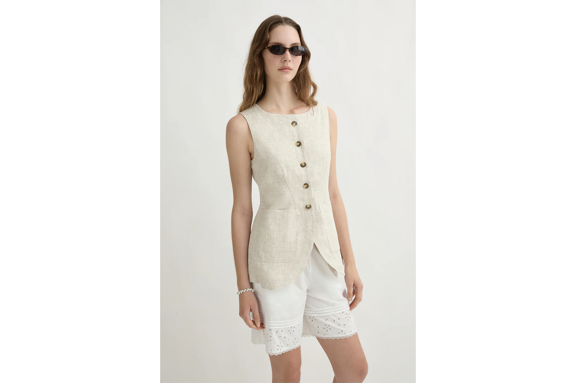 A female model wearing a beige linen sleeveless button-up top with a split hem, white embroidered shorts, sunglasses, and a bracelet.