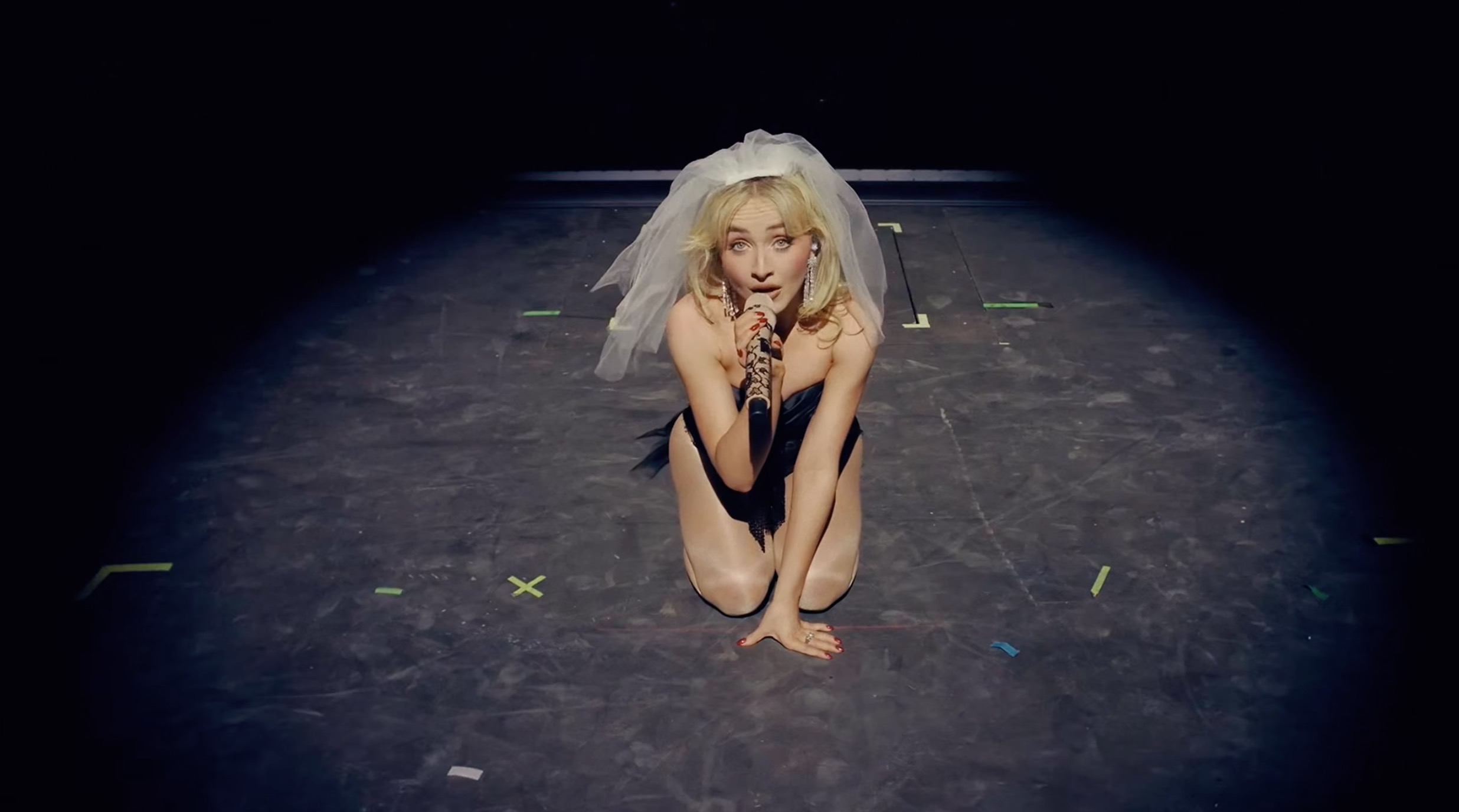 Sabrina Carpenter kneeling and singing into a microphone on stage, wearing a veil and black outfit.