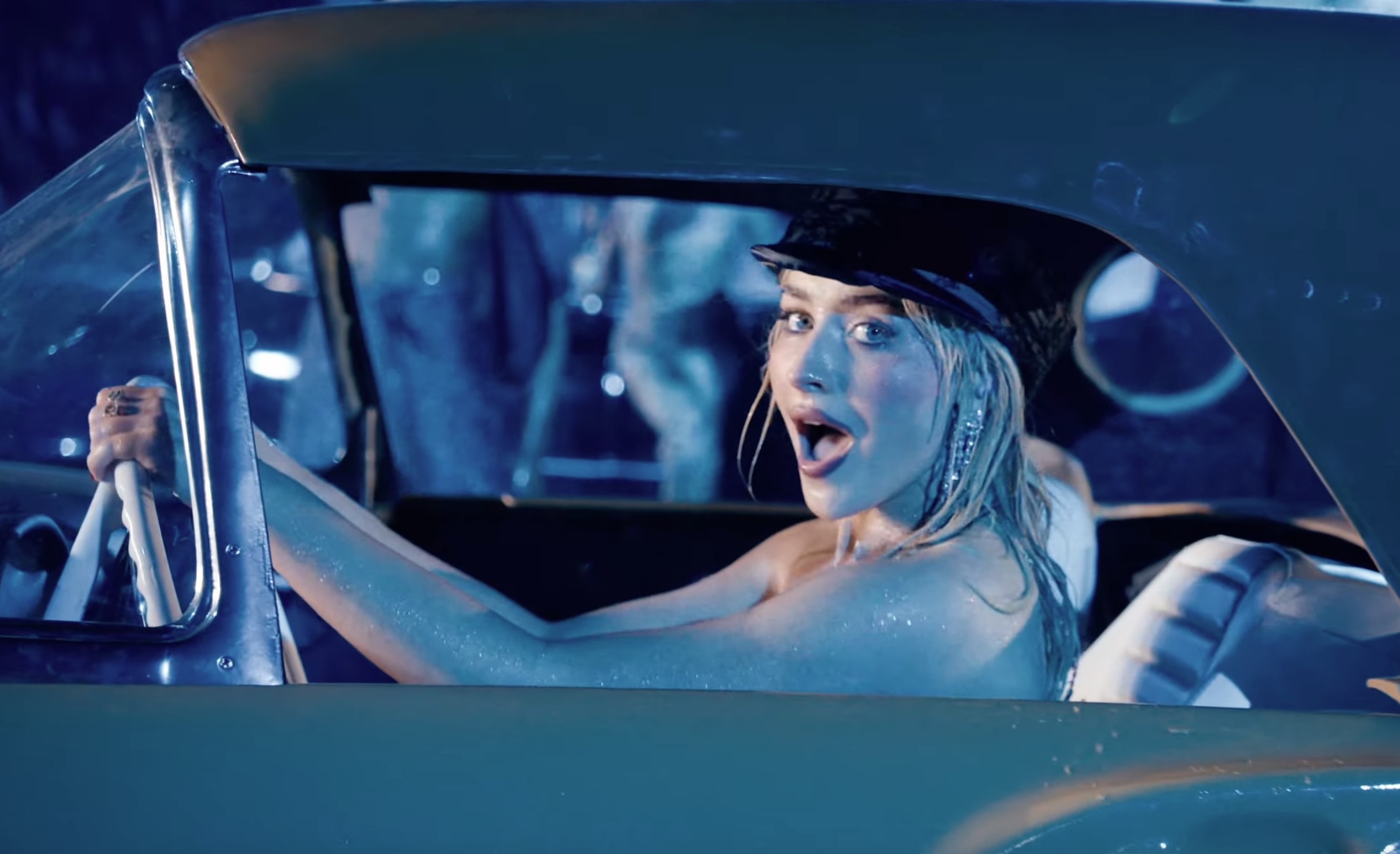 Sabrina Carpenter looking surprised while driving a vintage car.