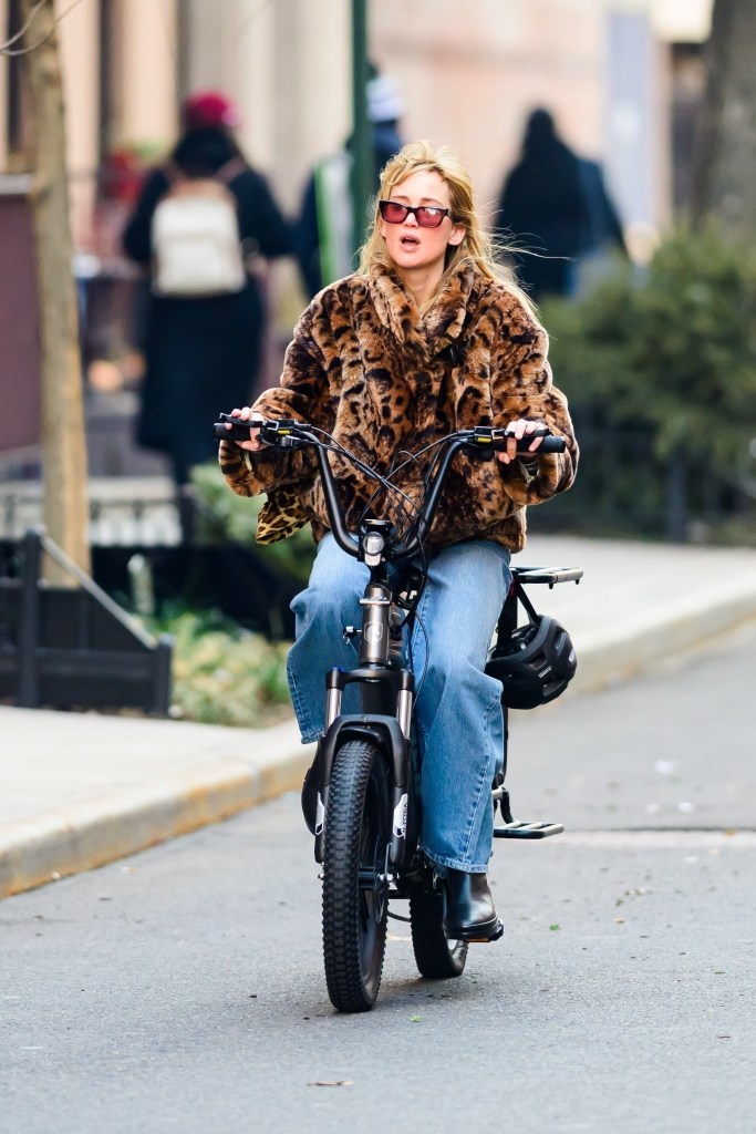 Jennifer Lawrence riding a Hovsco e-bike in New York, wearing a Jacquemus leopard print coat, Garrett Leight Woz sunglasses, La Ligne jeans, and The Row shoes.