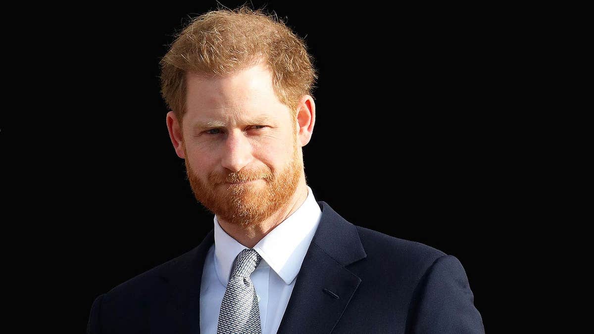 Prince Harry looking serious in a suit against a black background.