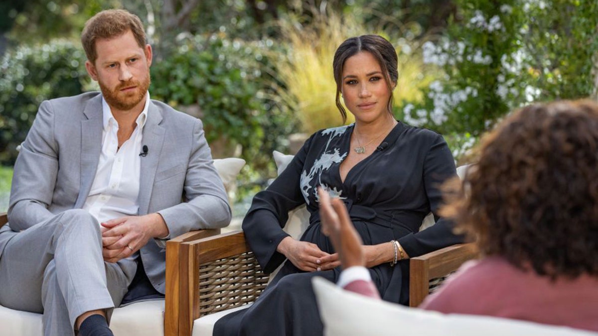 Prince Harry in grey suit and Meghan Markle in blue dress sitting across from Oprah Winfrey during interview