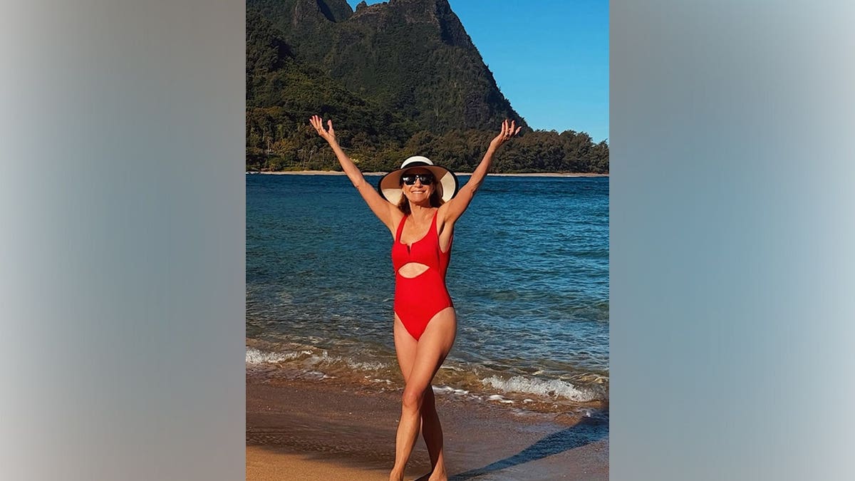 Jane Seymour wearing a red one-piece swimsuit and sun hat walking on a beach with mountains and ocean in the background.