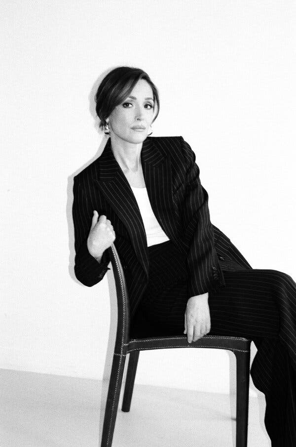 Rose Byrne sits in a chair, turning at the waist to face the camera.