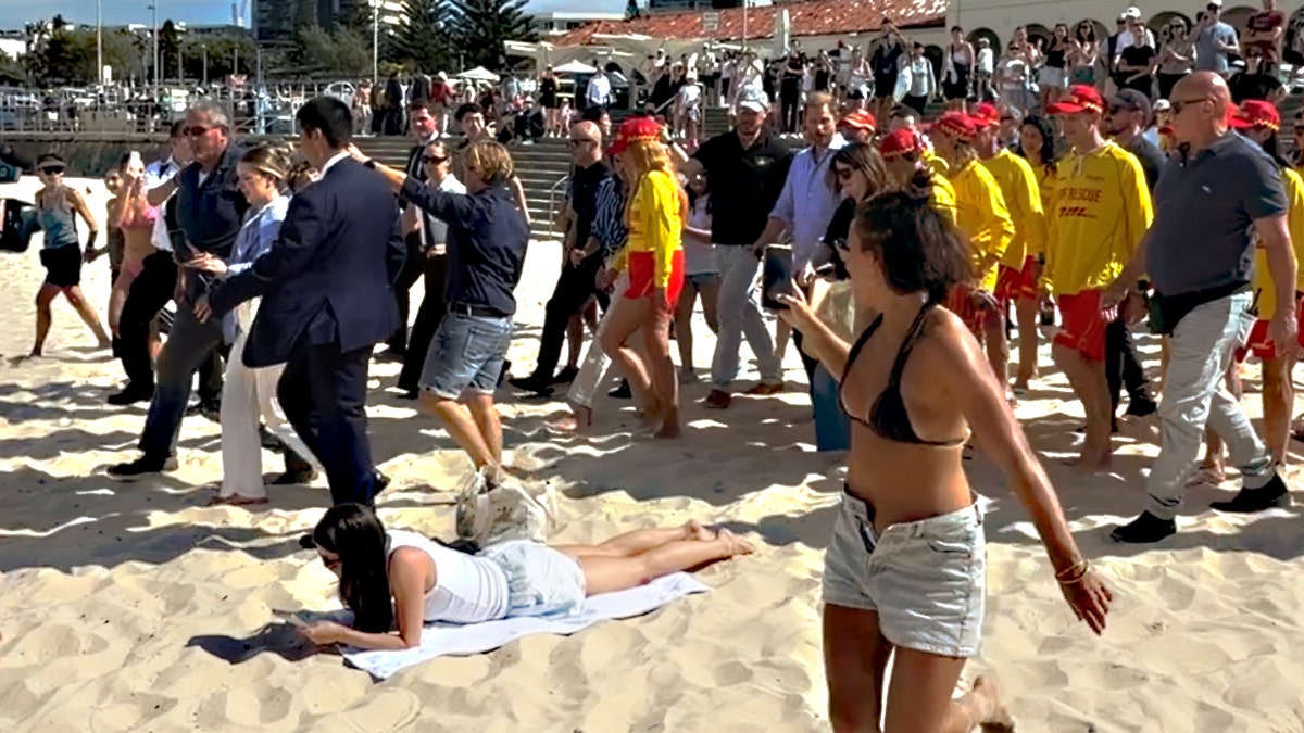 Prince Harry and Meghan Markle walking across a crowded beach in Sydney past a sunbather lying on the sand.