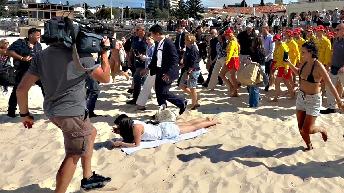 Prince Harry and Meghan Markle walking across a crowded beach in Sydney past a sunbather lying on the sand.
