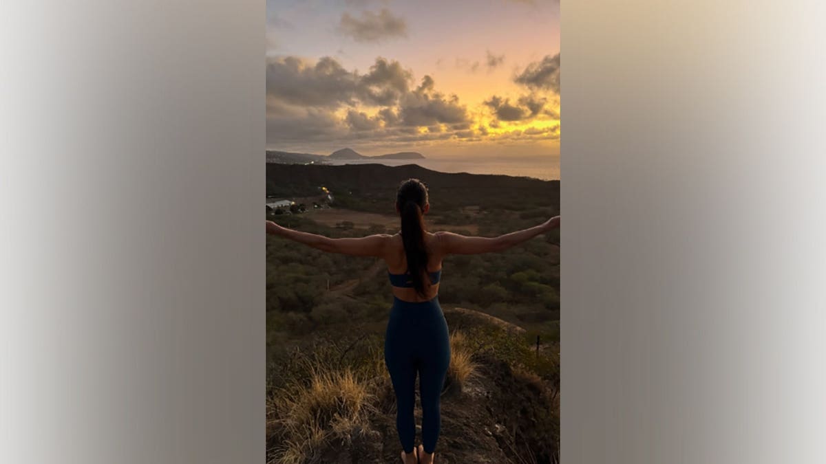 Nicole Scherzinger standing on a ridge with her back to the camera and arms spread wide overlooking a lush landscape at sunset.