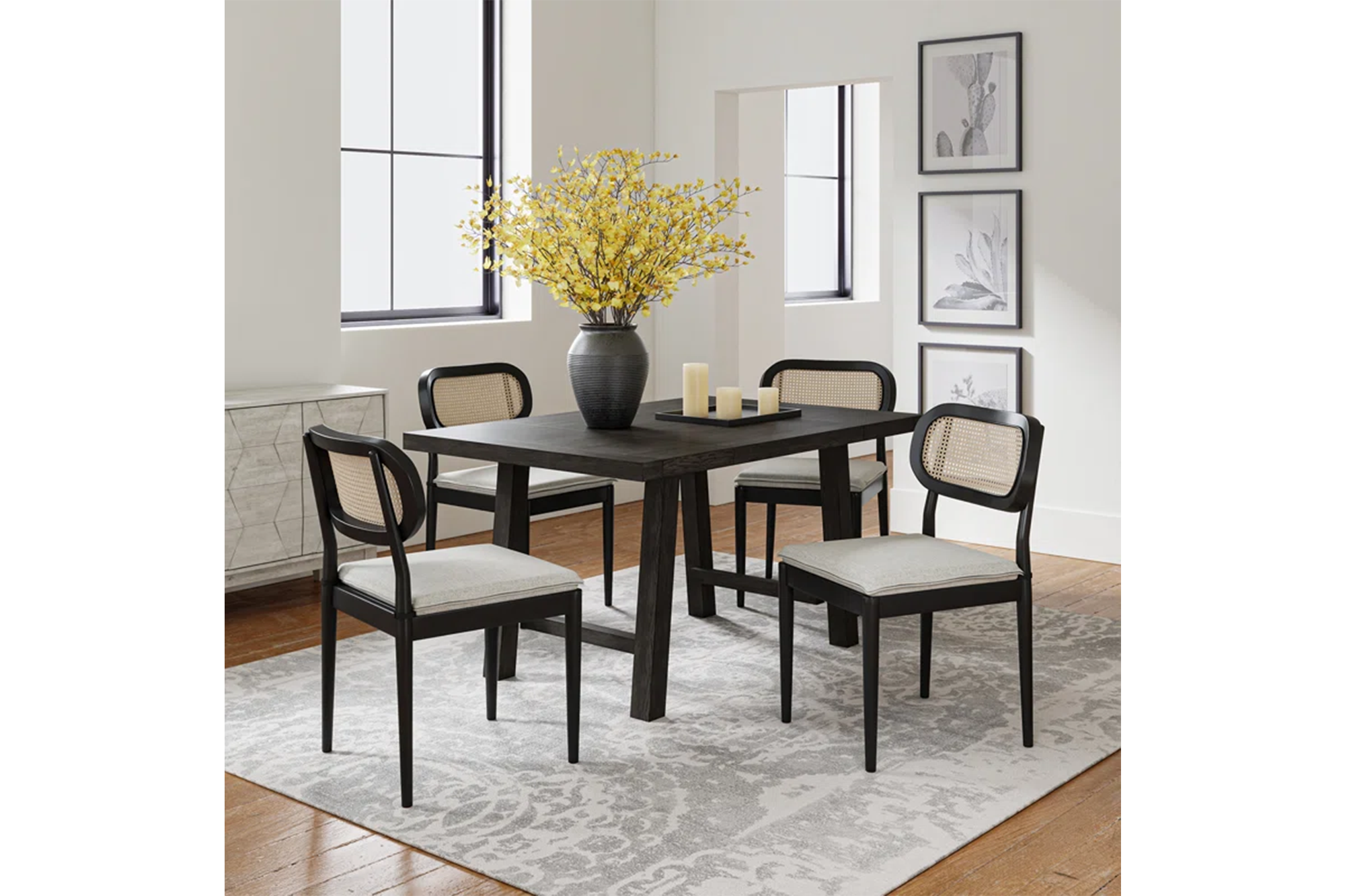 A black wooden dining table with four black chairs with cane backs and light gray cushioned seats on a gray and white patterned rug.