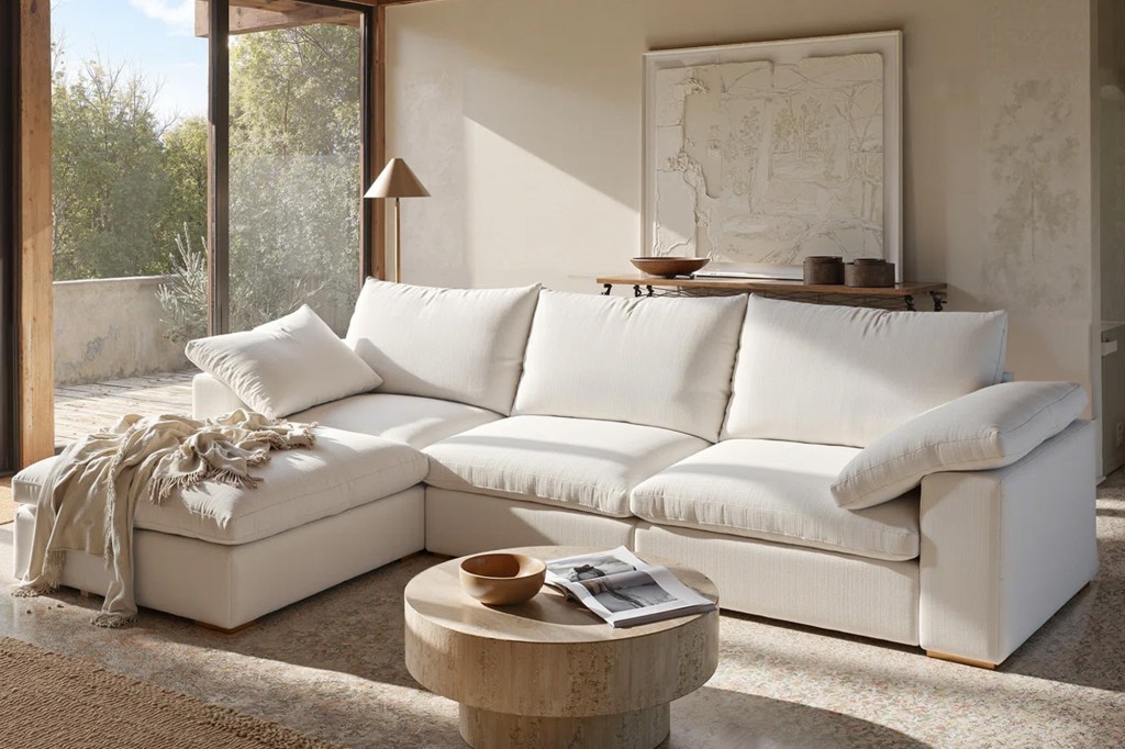 Living room with a large L-shaped white sectional sofa, round wooden coffee table, and large window overlooking trees.