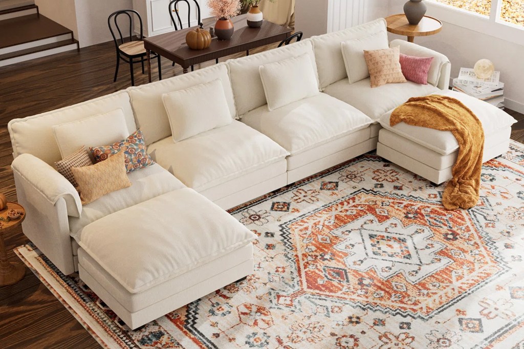 An L-shaped off-white modular sectional sofa with various throw pillows and a burnt orange patterned rug.