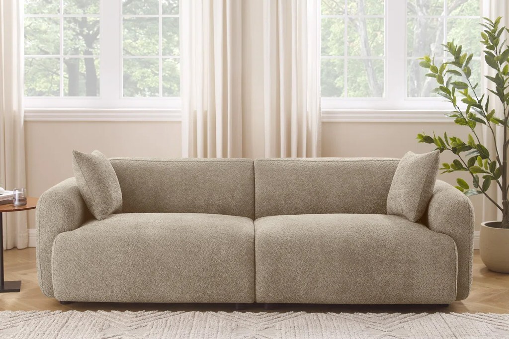 A beige textured sofa with two pillows in a living room with large windows, curtains, and a plant.
