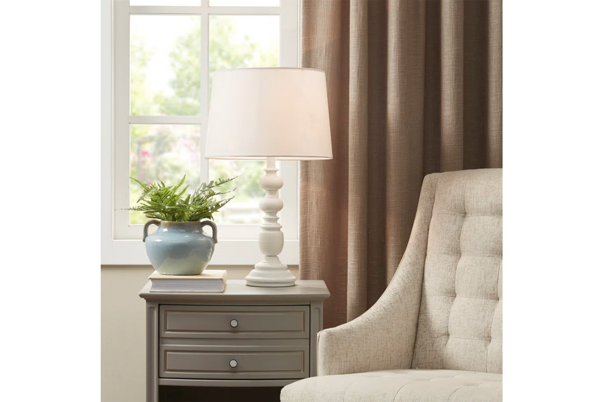 A white lamp, potted plant, and book on a gray nightstand next to a beige armchair.