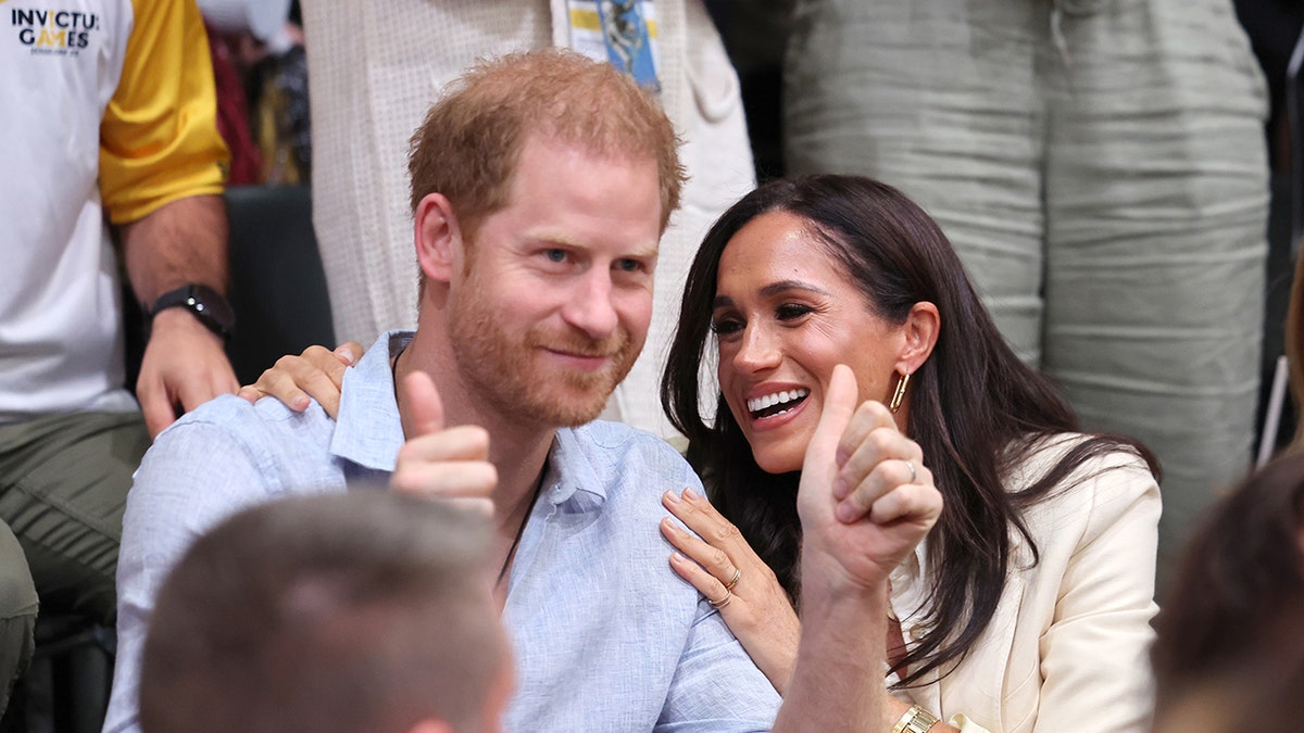 Prince Harry giving thumbs up with Meghan Markle smiling and leaning on him at a volleyball match